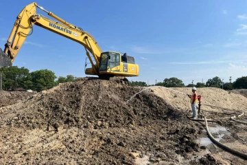 A worker spraying water on a pile of dirt. On the top of the dirt there is a yellow excavator